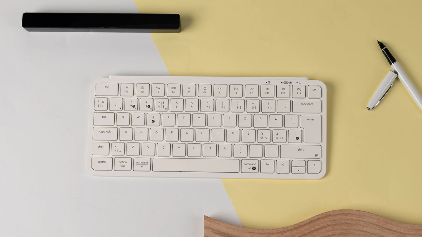 Eine weiße Tastatur, ein Stift und ein schwarzer Balken liegen auf gelbem und weißem Hintergrund. Unten ist ein Holzgegenstand.