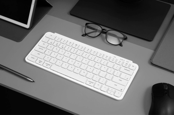 Weiße Tastatur, Brille, Stift, Maus und Tablet auf grauer Oberfläche. Verschwommener Hintergrund, Arbeitsplatz.