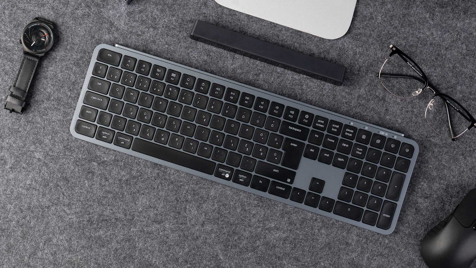 Draufsicht mit Tastatur, Uhr, Brille und Maus auf grauer Oberfläche. Die Tastatur steht im Fokus.