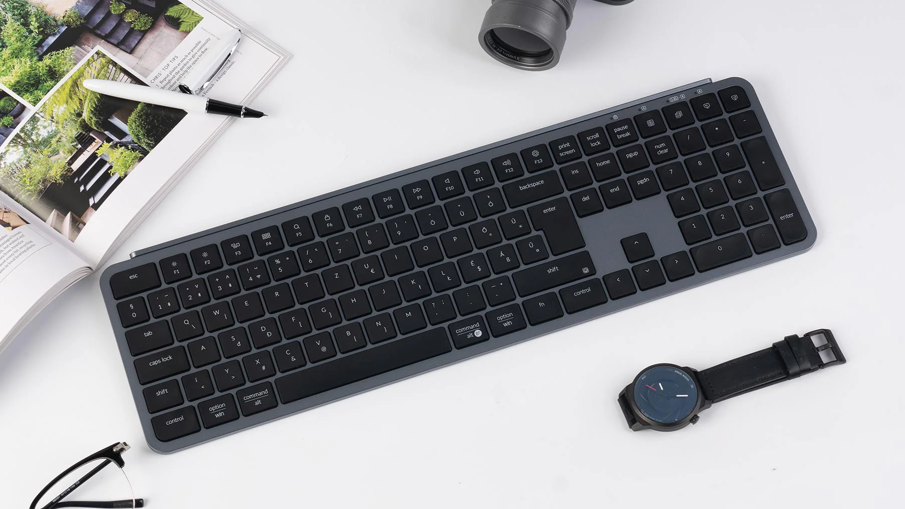 Eine Tastatur, Uhr, Stift, Brille und Zeitschrift liegen auf einer weißen Oberfläche. Die Tastatur ist grau. Die Uhr hat ein schwarzes Armband.