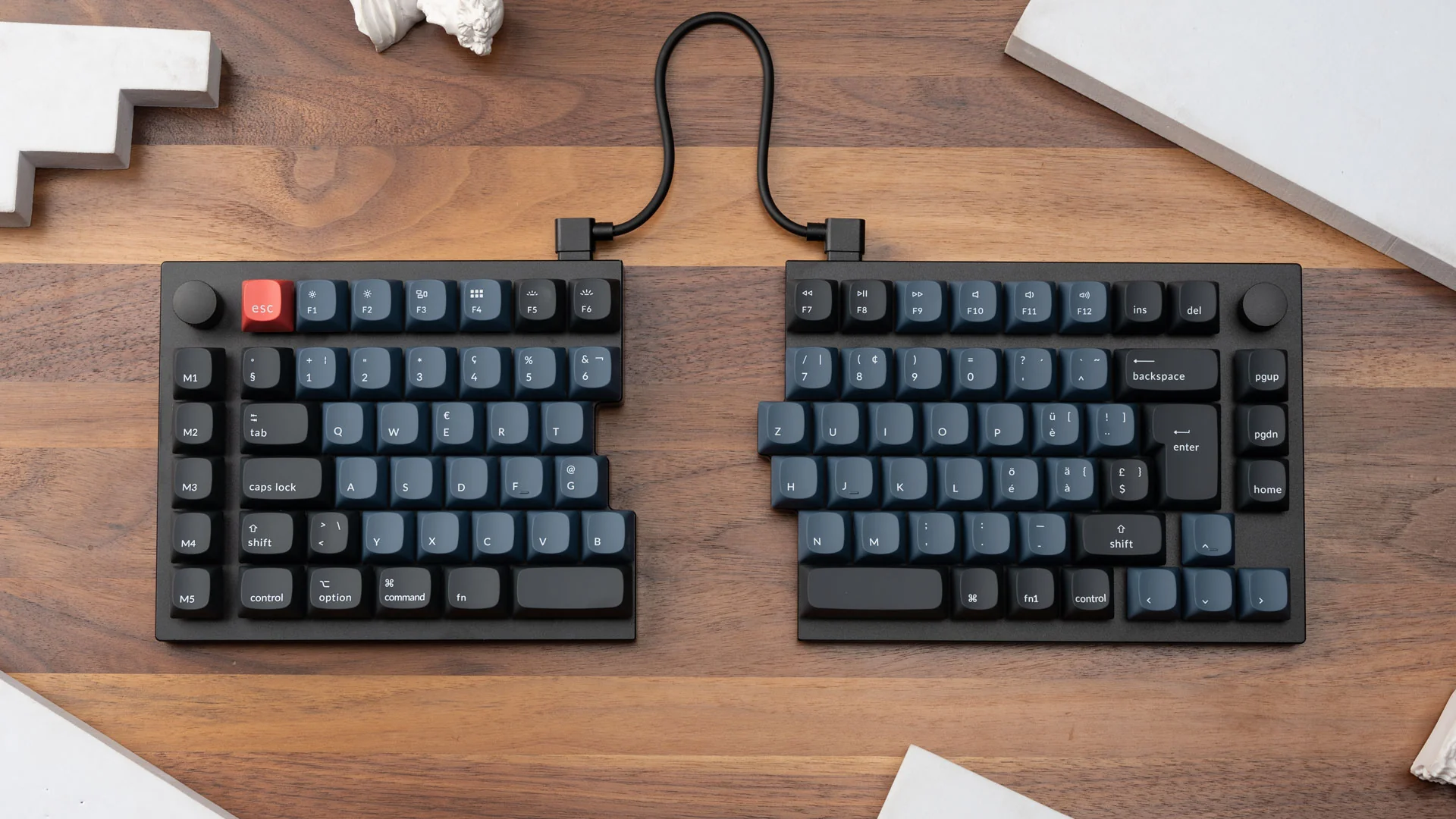 Geteilte Tastatur mit schwarzen Tasten und blauen Beschriftungen. Rote ESC-Taste. Verbunden durch ein schwarzes Kabel. Holz-Tisch-Hintergrund.