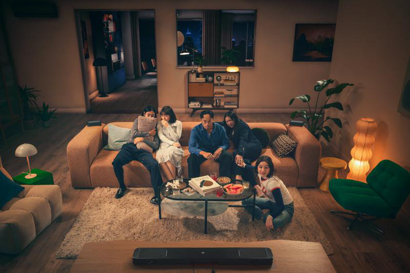 Eine Familie sitzt auf einem Sofa und schaut etwas an. Ein Glastisch mit Essen steht davor. Eine Soundbar ist im Vordergrund.