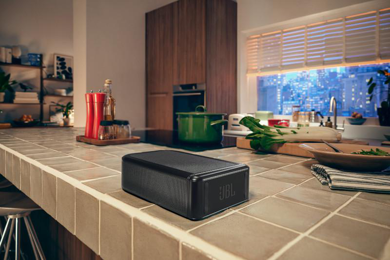 Ein schwarzer JBL-Lautsprecher steht auf einer Küchentheke. Weitere Gegenstände sind ein Topf, Gemüse und ein Blick auf die Stadt.