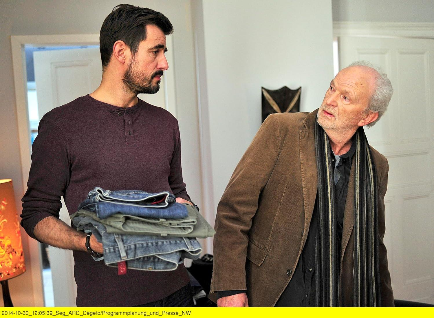 Ein Mann hält gefaltete Jeans und schaut einen anderen Mann in einer braunen Jacke an. Hintergrund: weiße Wände und Tür.