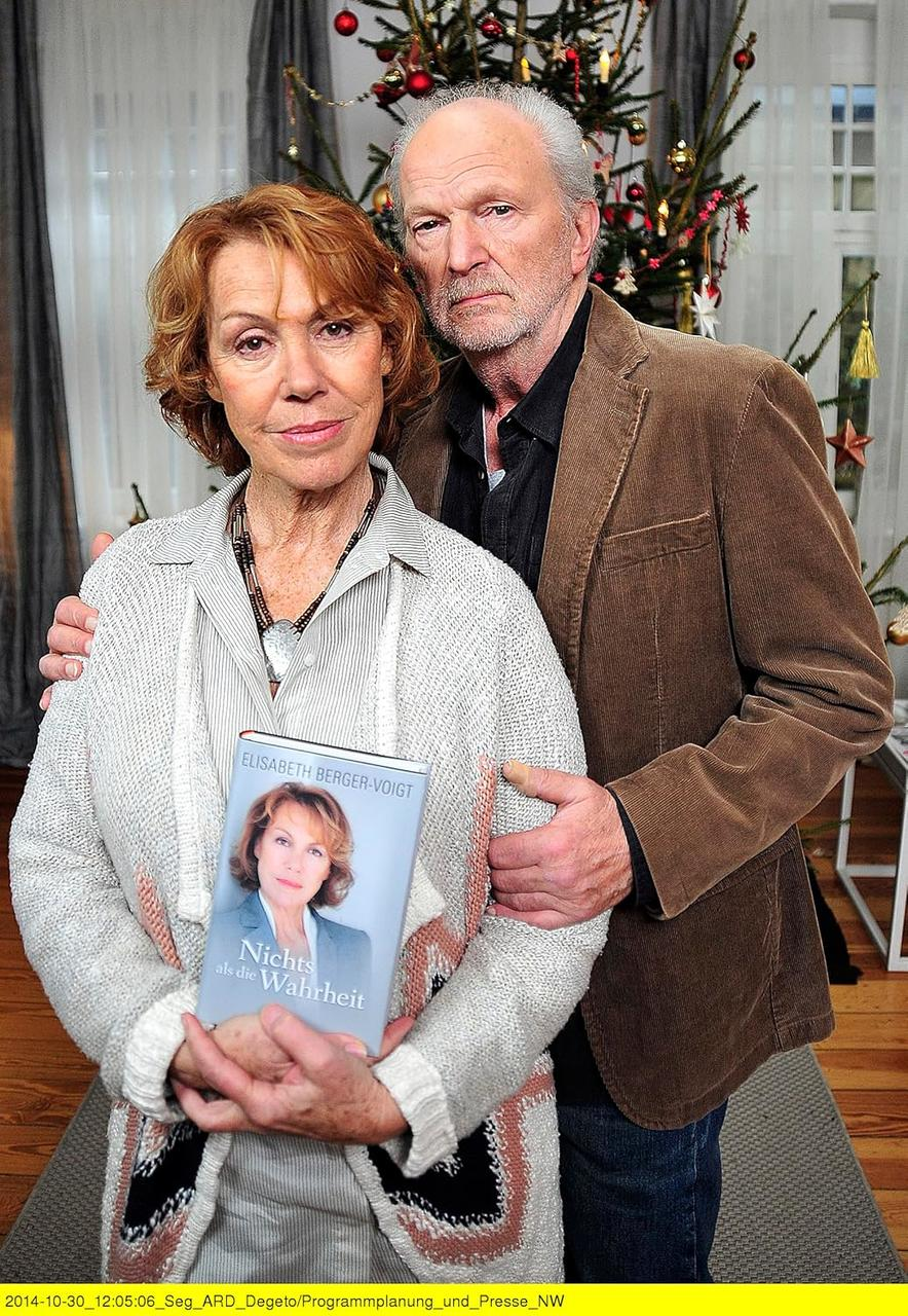 Ältere Frau und Mann posieren zusammen, die Frau hält ein Buch. Im Hintergrund ist ein Weihnachtsbaum zu sehen.