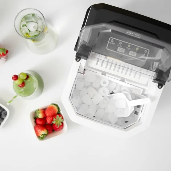 Maszyna do lodu jest otwarta, obok zielonego smoothie z owocami, szklanki wody z lodem i owocami oraz truskawkami.