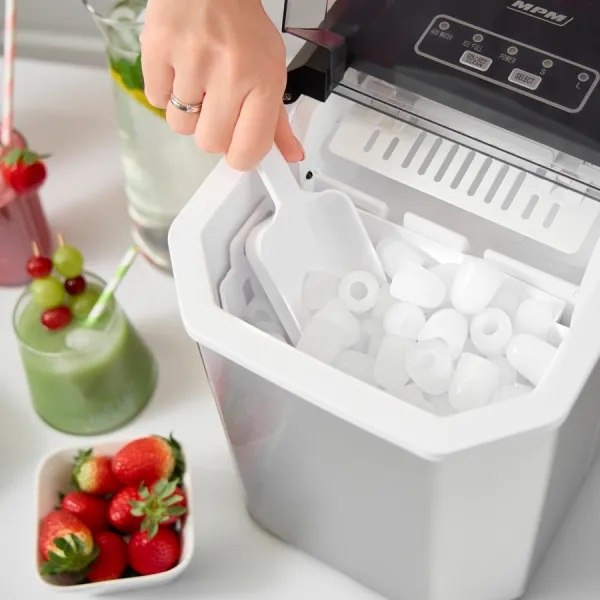 Osoba używa białej maszyny do lodu. Kostki lodu są widoczne. Truskawki i zielony koktajl na pierwszym planie.