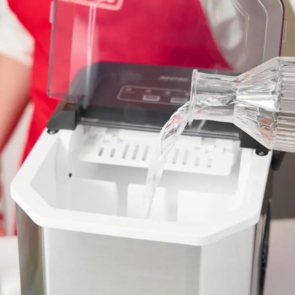 Woda wlewana do kostkarki do lodu. Kostkarka jest biała i srebrna. W tle osoba ubrana na czerwono.