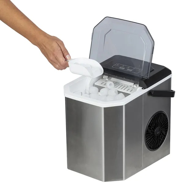 Ręka nabiera lód z kostkarki. Maszyna jest srebrna i czarna. Pokrywa jest otwarta. Kostki lodu są widoczne w środku.