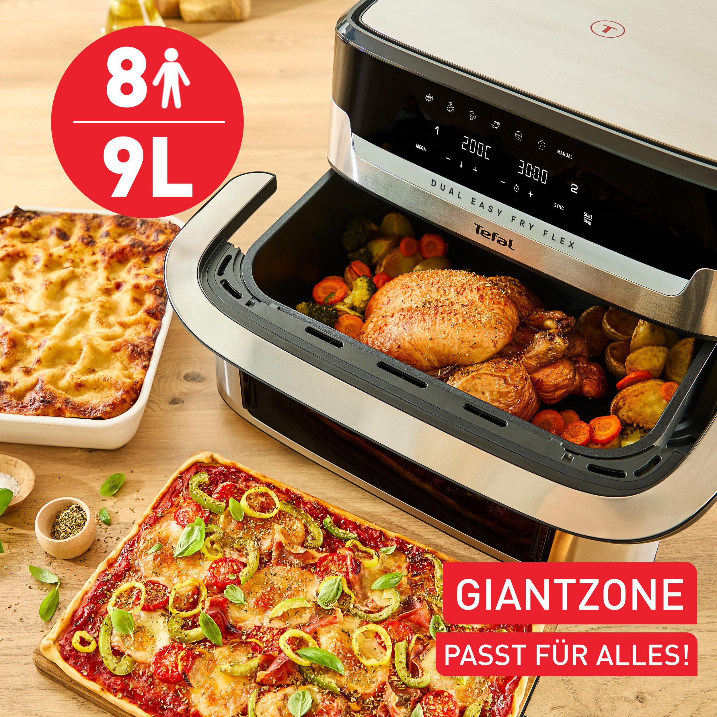 Ein Airfryer mit gekochtem Hähnchen und Gemüse sowie Pizza und Lasagne daneben auf einem Holztisch.