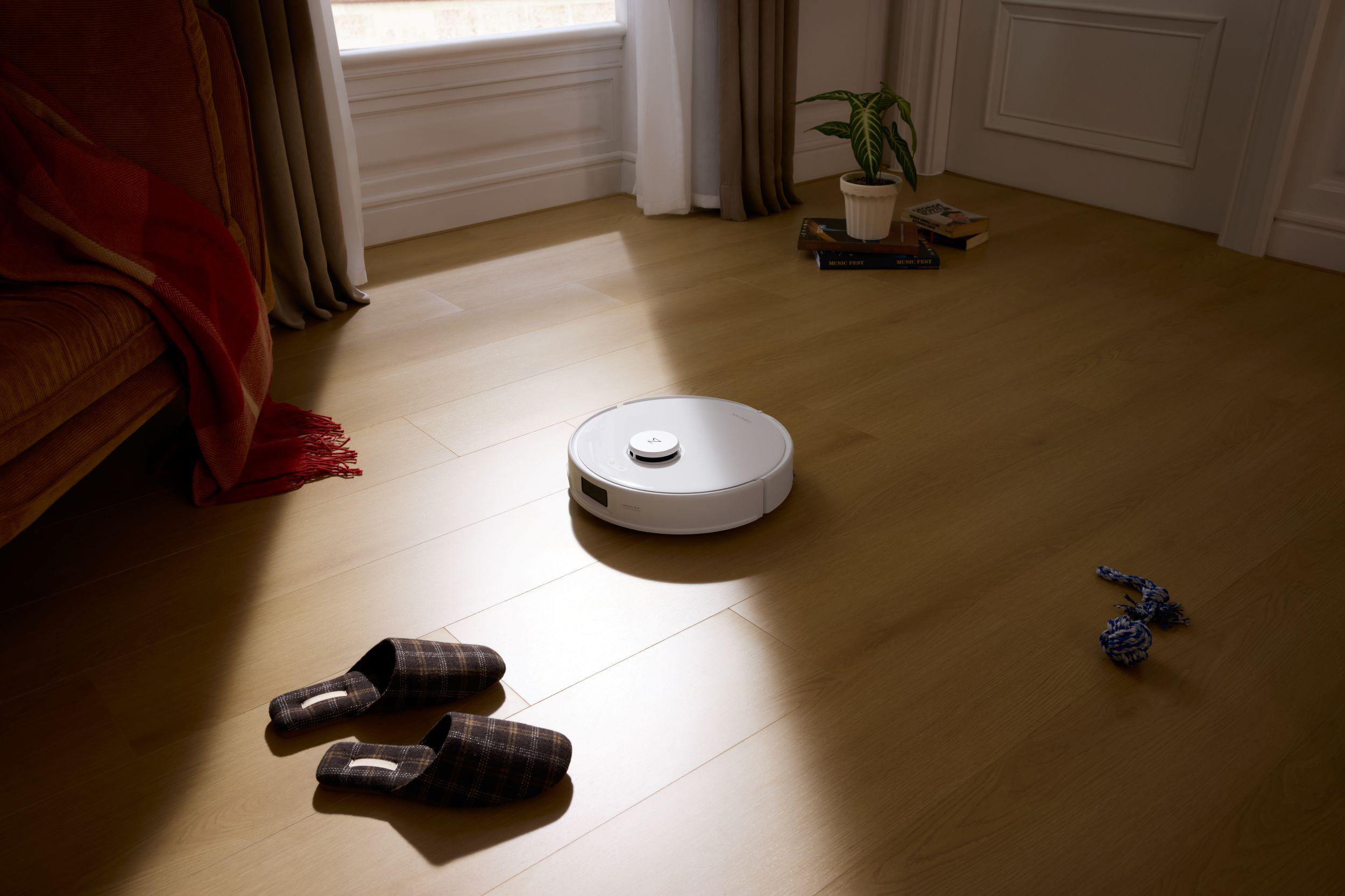 Ein weißer Roboter-Staubsauger auf Holzboden. Hausschuhe und ein Hundespielzeug sind in der Nähe.
