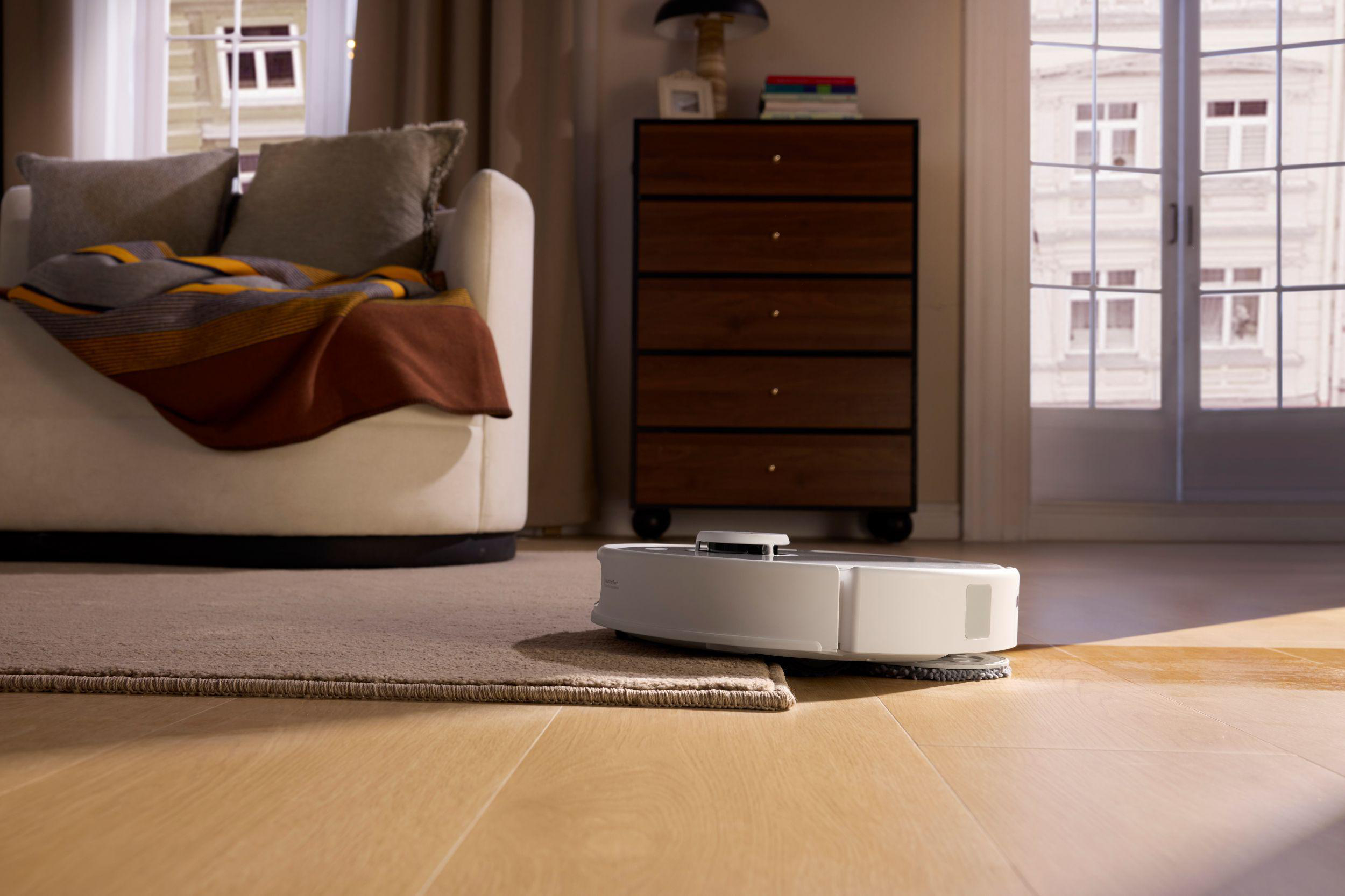 Ein weißer Roboterstaubsauger auf Holzboden neben einem Teppich. Im Hintergrund ein Sofa mit Kissen und Decke.