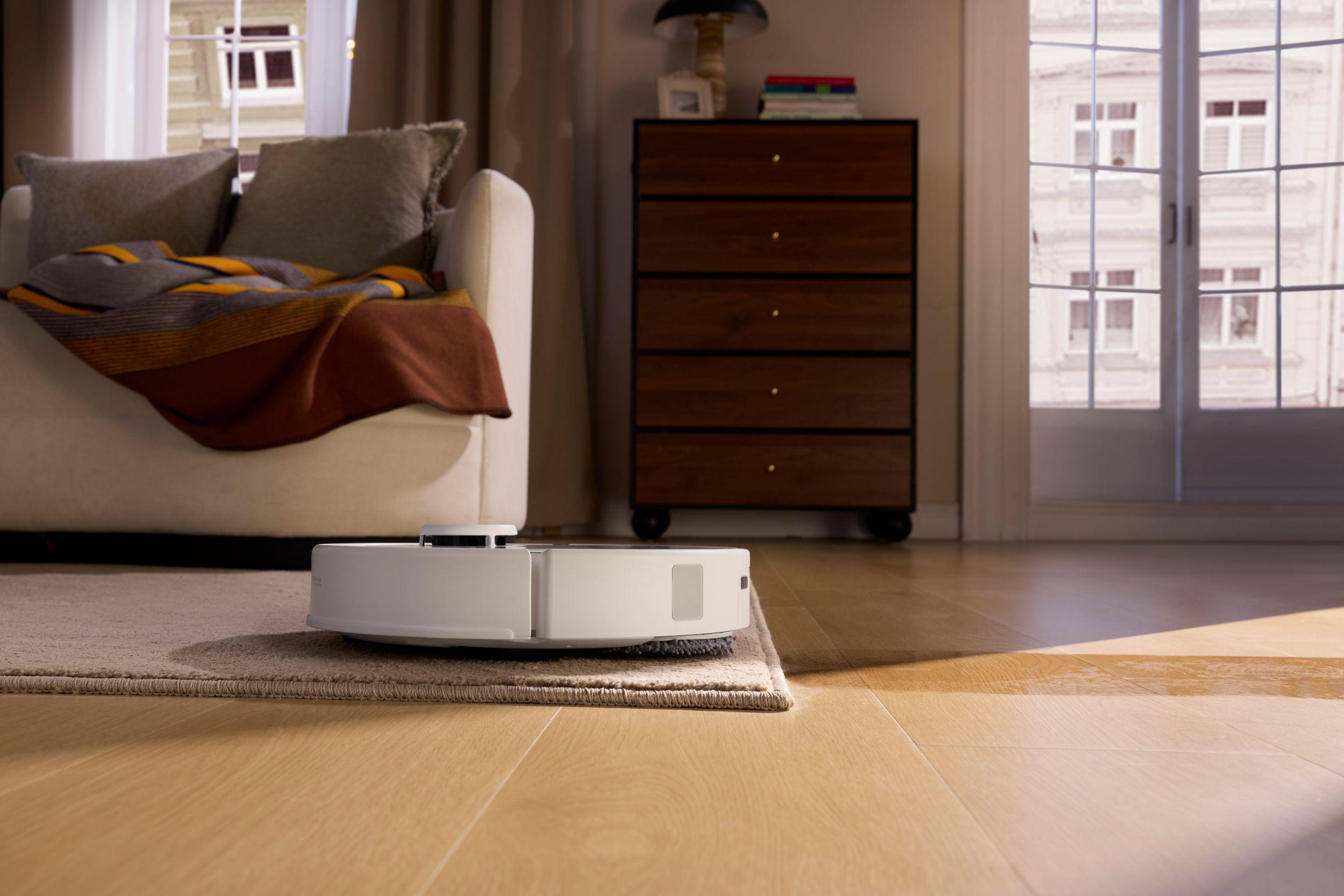 Ein weißer Roboterstaubsauger auf einem Teppich. Im Hintergrund ein Sofa mit Kissen und Decke. Holzboden.