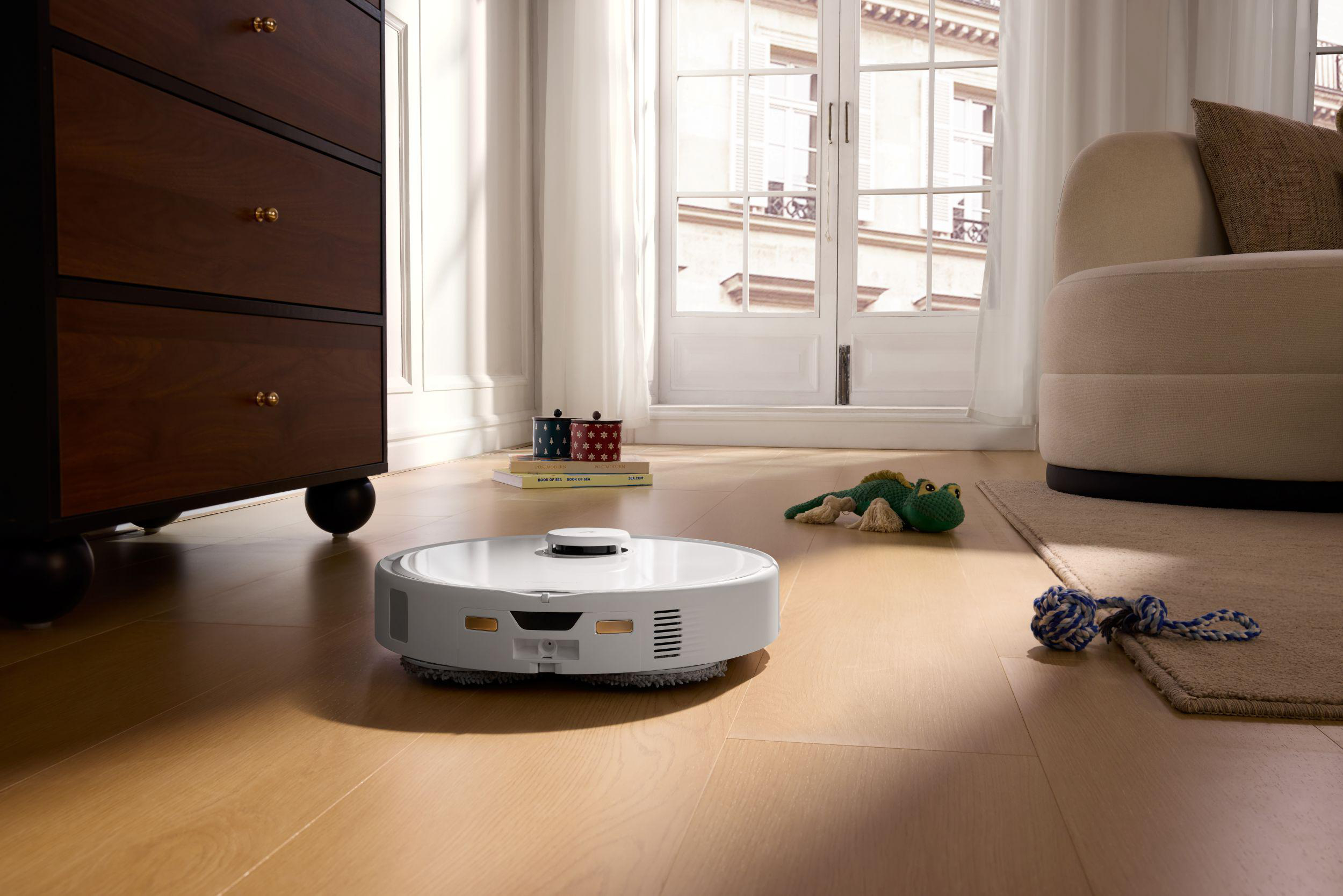 Ein weißer Roboterstaubsauger reinigt einen Raum mit Holzboden. Eine dunkelbraune Kommode und ein Sofa sind an den Seiten.