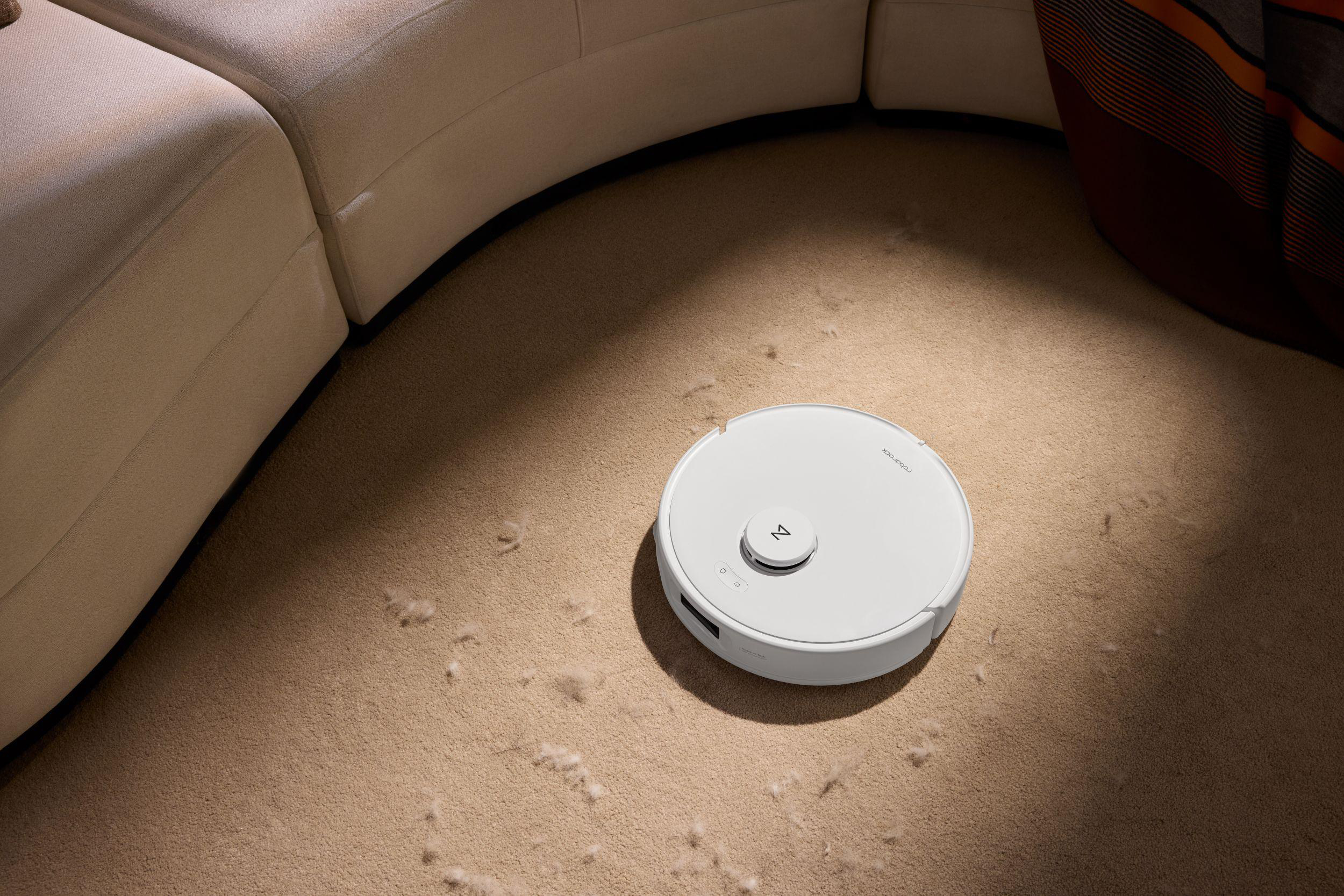 Ein weißer Roboterstaubsauger reinigt einen Teppich mit Tierhaaren. Links ein gebogenes Sofa, rechts eine Decke.