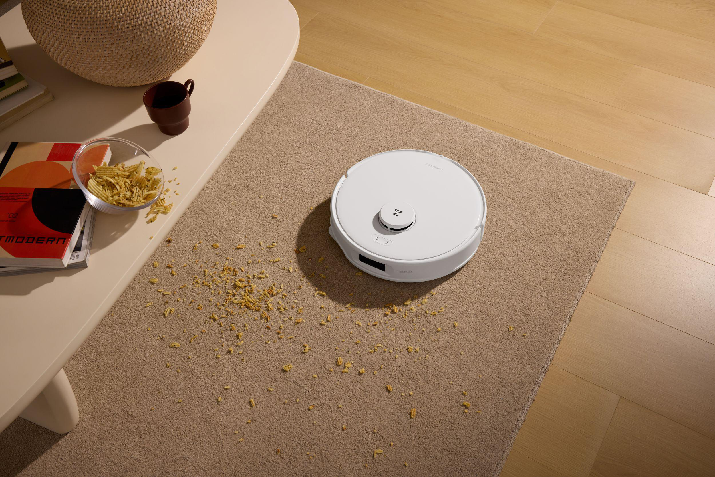 Ein weißer Roboterstaubsauger auf einem beigen Teppich. Chips und eine Tasse stehen auf einem Tisch. Holzboden im Hintergrund.