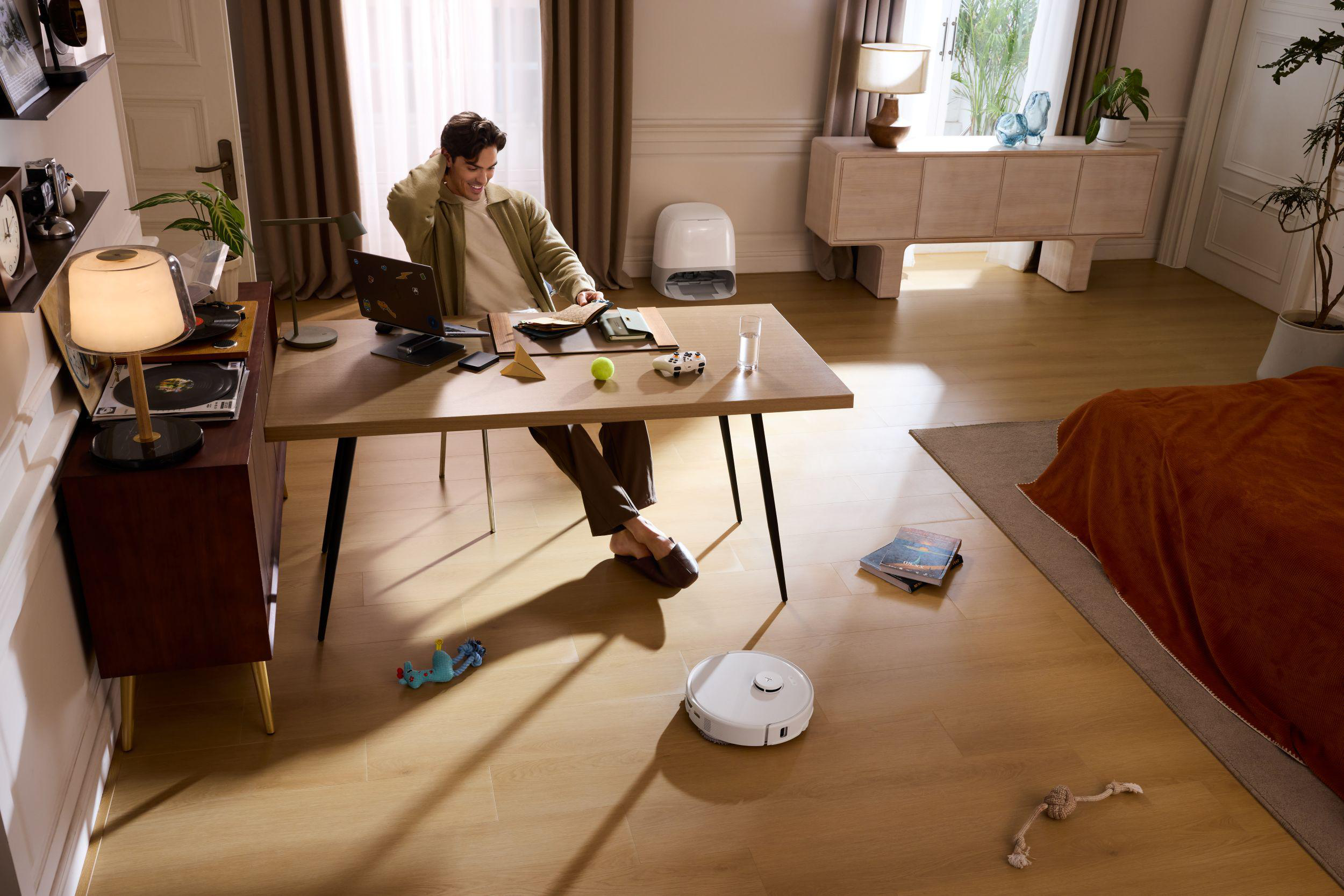 Mann am Schreibtisch, Laptop offen. Roomba auf dem Boden, Tennisball und Spielzeug. Holzfußboden und Möbel.