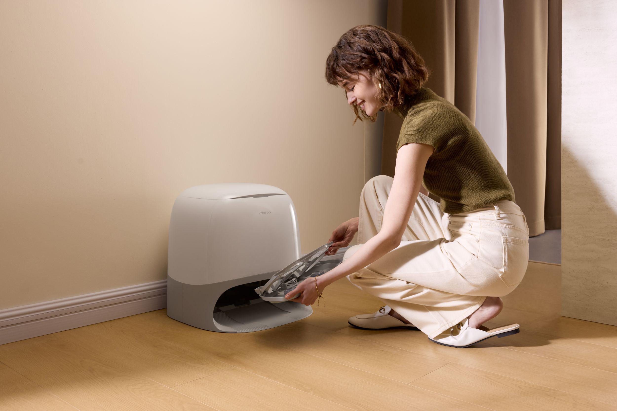 Frau in grünem Shirt hockt neben einer grauen Katzentoilette und hält ein Tablett. Beige Wand und Holzboden.
