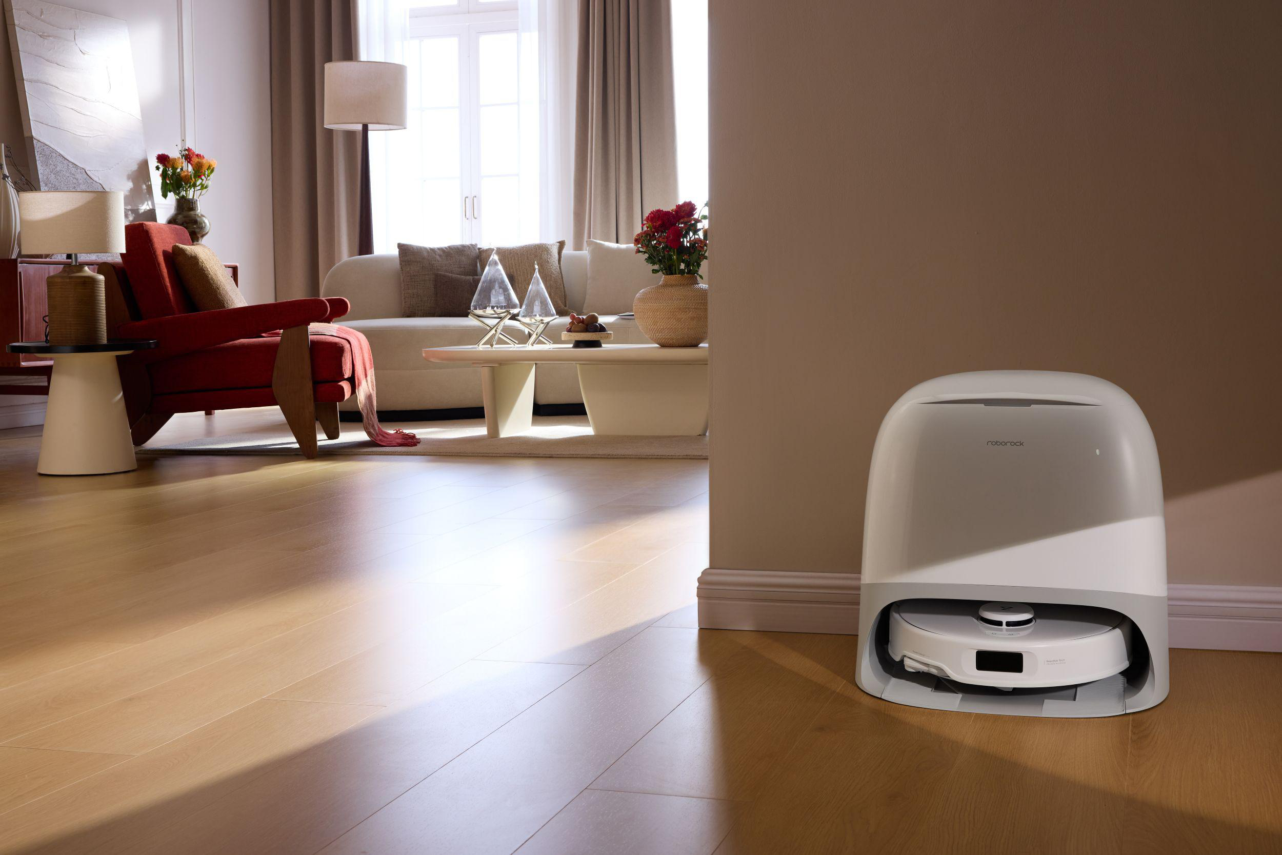 Ein modernes Wohnzimmer mit einem Roboter-Staubsauger im Vordergrund. Roter Stuhl, weißes Sofa und Blumen sind sichtbar.