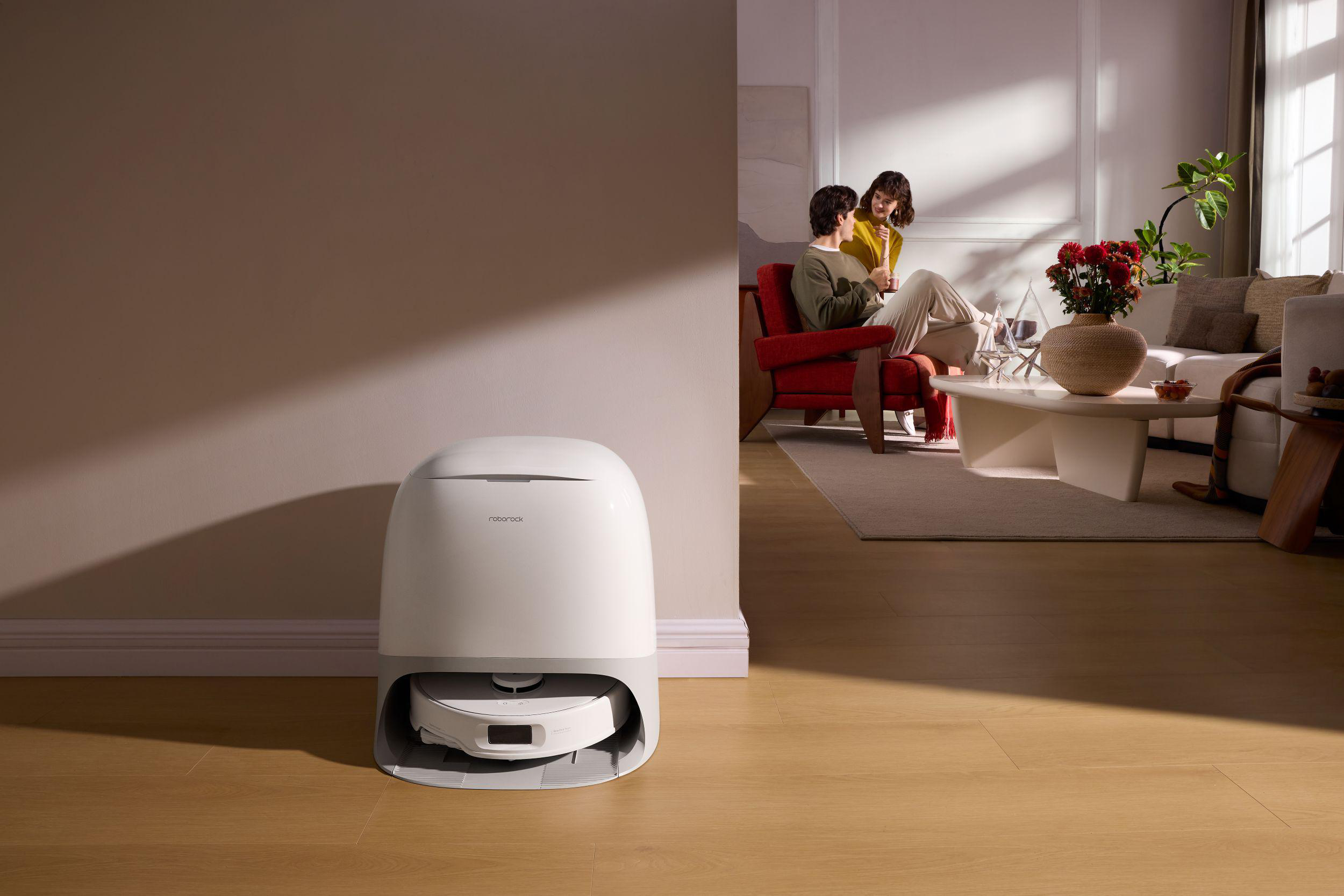 Ein weiß-grauer Roboter-Staubsauger im Vordergrund. Im Hintergrund sitzt ein Paar in einem Wohnzimmer.