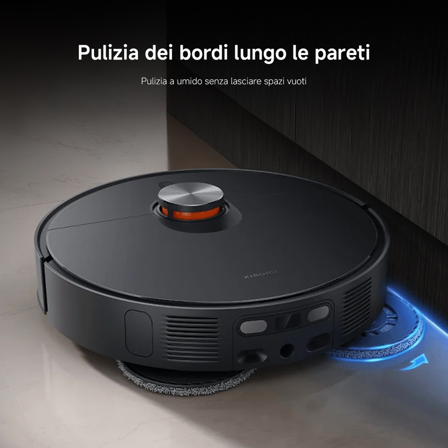 Un aspirapolvere robotico nero pulisce un pavimento, vicino a un muro scuro. L'aspirapolvere ha una forma rotonda e la parte superiore arancione.