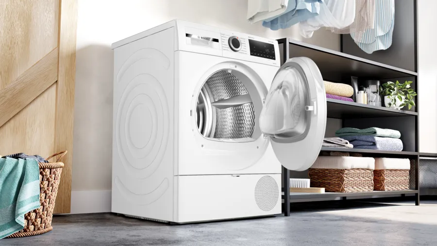 Un sèche-linge blanc est ouvert, révélant l'intérieur. Un panier à linge est à gauche, et des étagères avec des serviettes à droite.