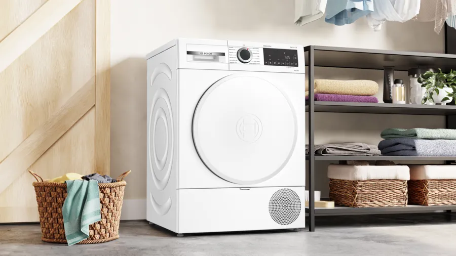 Un sèche-linge Bosch blanc se trouve près d'une porte en bois et d'une étagère avec des serviettes pliées et des paniers. Un panier à linge est devant.