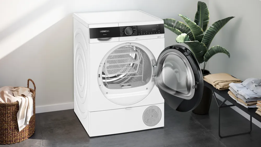 Ein weißer Siemens-Trockner mit geöffneter Tür. Links ein Wäschekorb. Rechts eine Pflanze und zusammengelegte Kleidung.