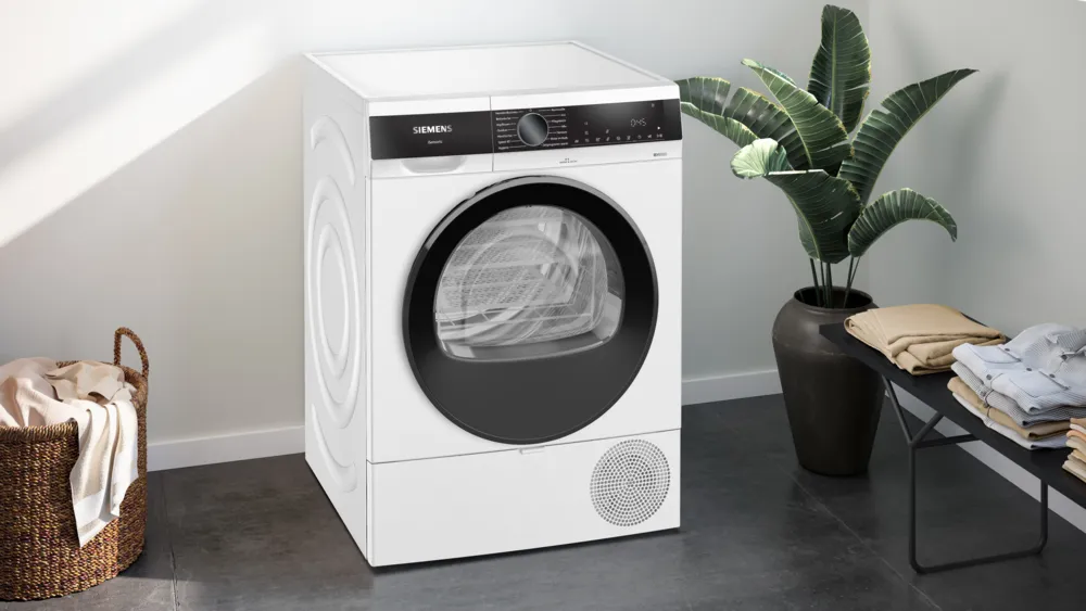 Ein weißer Siemens-Trockner steht neben einer Topfpflanze. Kleidung liegt auf einem schwarzen Tisch und in einem Weidenkorb.