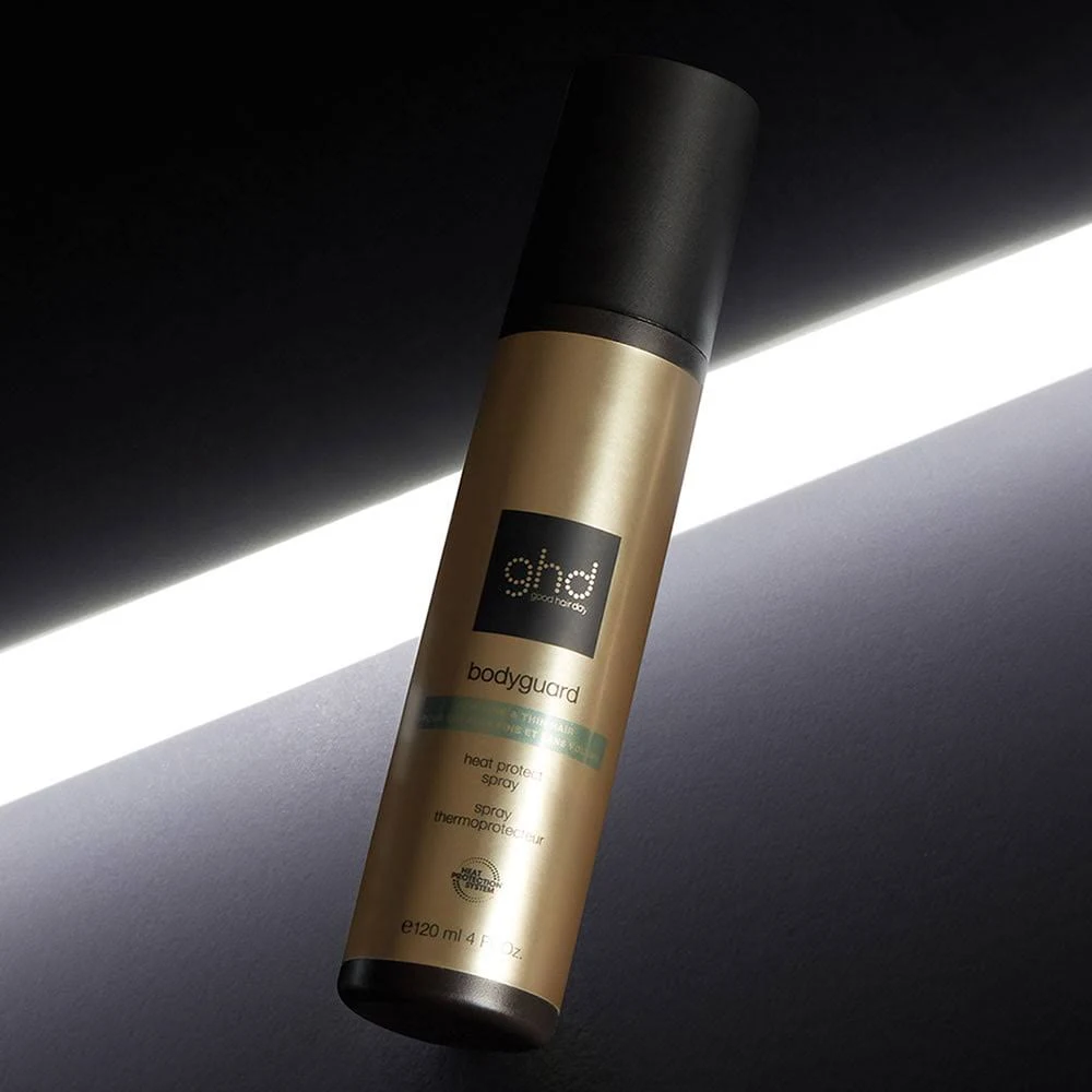 Un frasco dorado de spray protector térmico ghd Bodyguard con tapa negra, sobre un fondo oscuro con una franja de luz blanca.