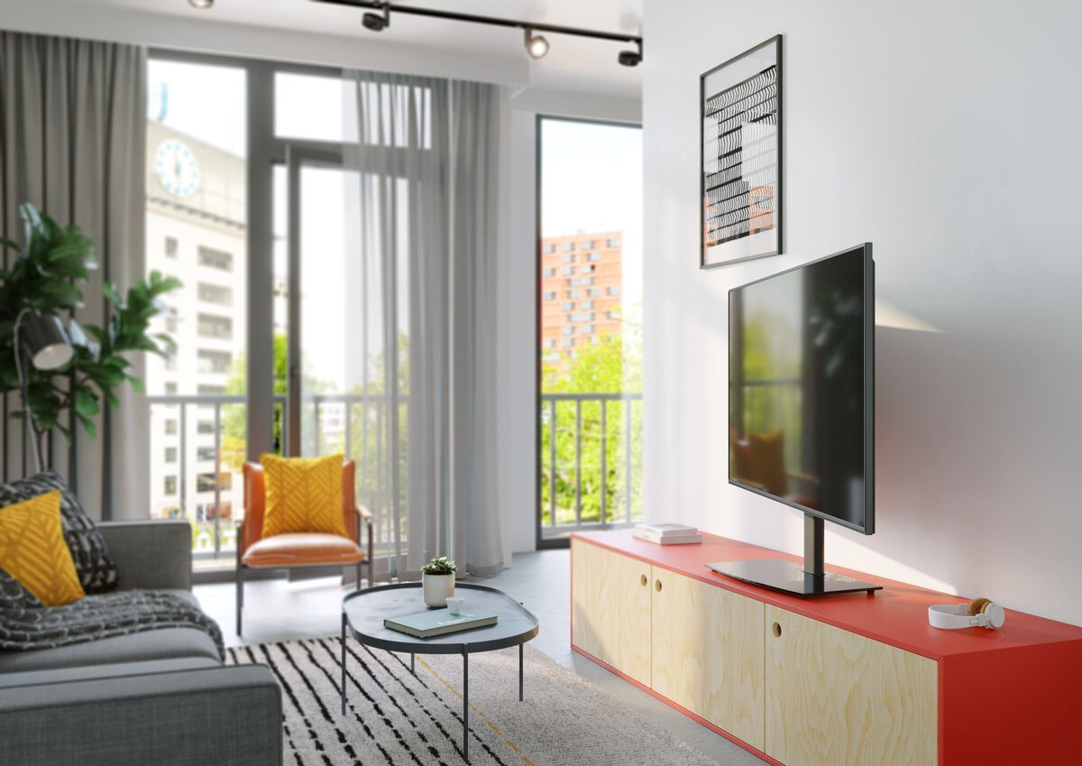 Una sala de estar con televisor en un mueble rojo, un sofá, una silla y una mesa de centro. Se ve un edificio por la ventana.