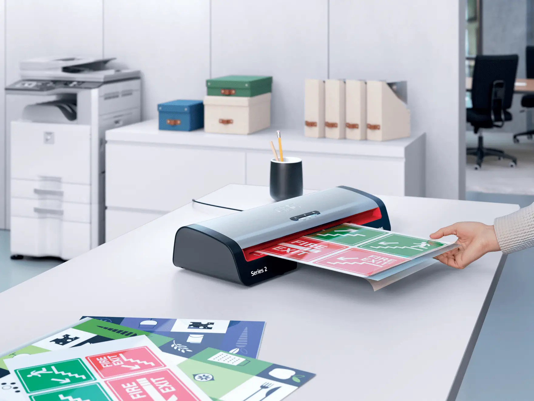 Egy személy tűzoltási kijárat táblákat laminál. Egy fehér irodai asztalon egy laminálógép, táblák és egy csésze ceruzákkal. További irodaszerek láthatók.