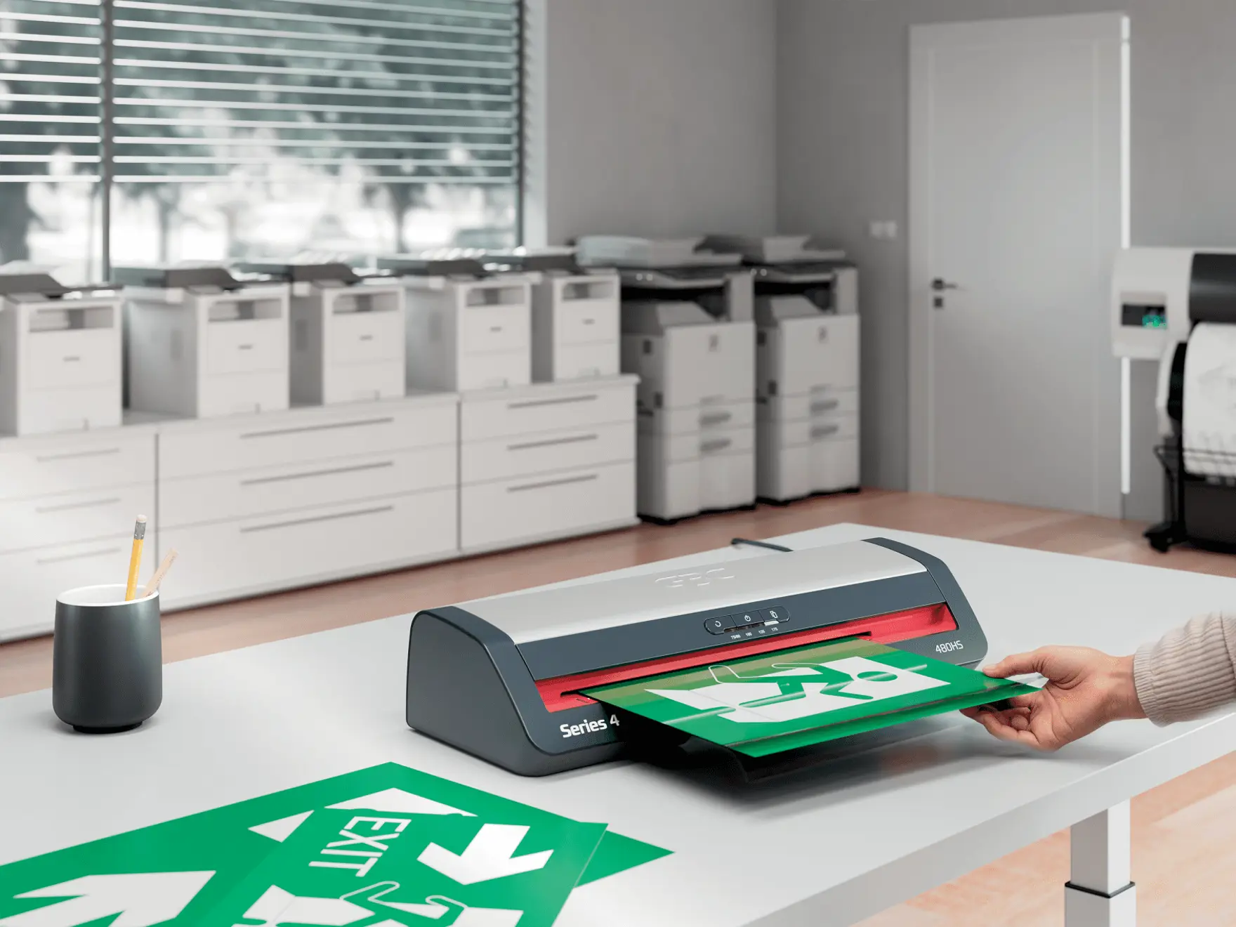 Egy személy egy zöld "KIJÁRAT" táblát laminál. Egy laminálógép és irodai berendezések egy fehér asztalon. A háttérben redőnyök és nyomtatók láthatók.