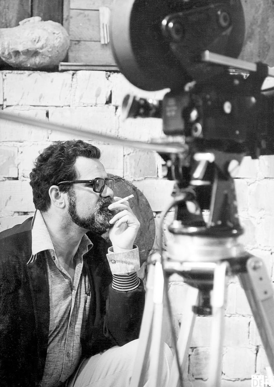 Mann mit Brille raucht Zigarette, sitzt neben Kamera. Ziegelwand im Hintergrund, Filmrolle im Hintergrund.