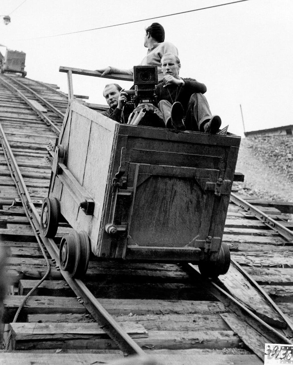 Männer fahren in einem Wagen auf Schienen und filmen. Ein Mann hält eine Kamera. Ein weiterer Wagen ist weiter oben am Hang.