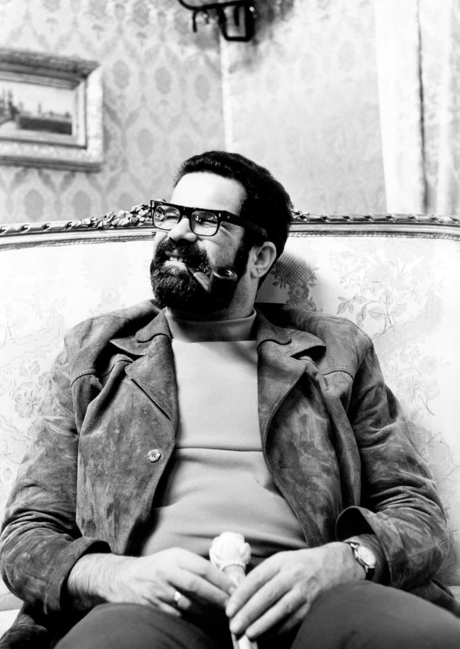 Ein Mann mit Brille und Bart sitzt auf einer floralen Couch und raucht eine Pfeife. Er hält einen Gegenstand in den Händen.