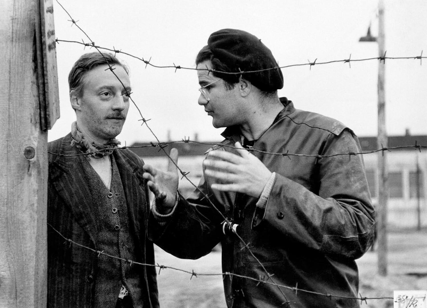 Zwei Männer hinter Stacheldraht, einer mit Baskenmütze, gestikuliert. Der andere, mit Weste, blickt besorgt.