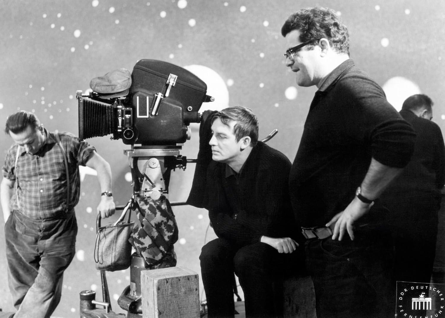 Männer um eine Filmkamera. Ein Mann blickt in die Kamera, ein anderer steht daneben. Der Hintergrund hat ein Muster aus weißen Punkten.