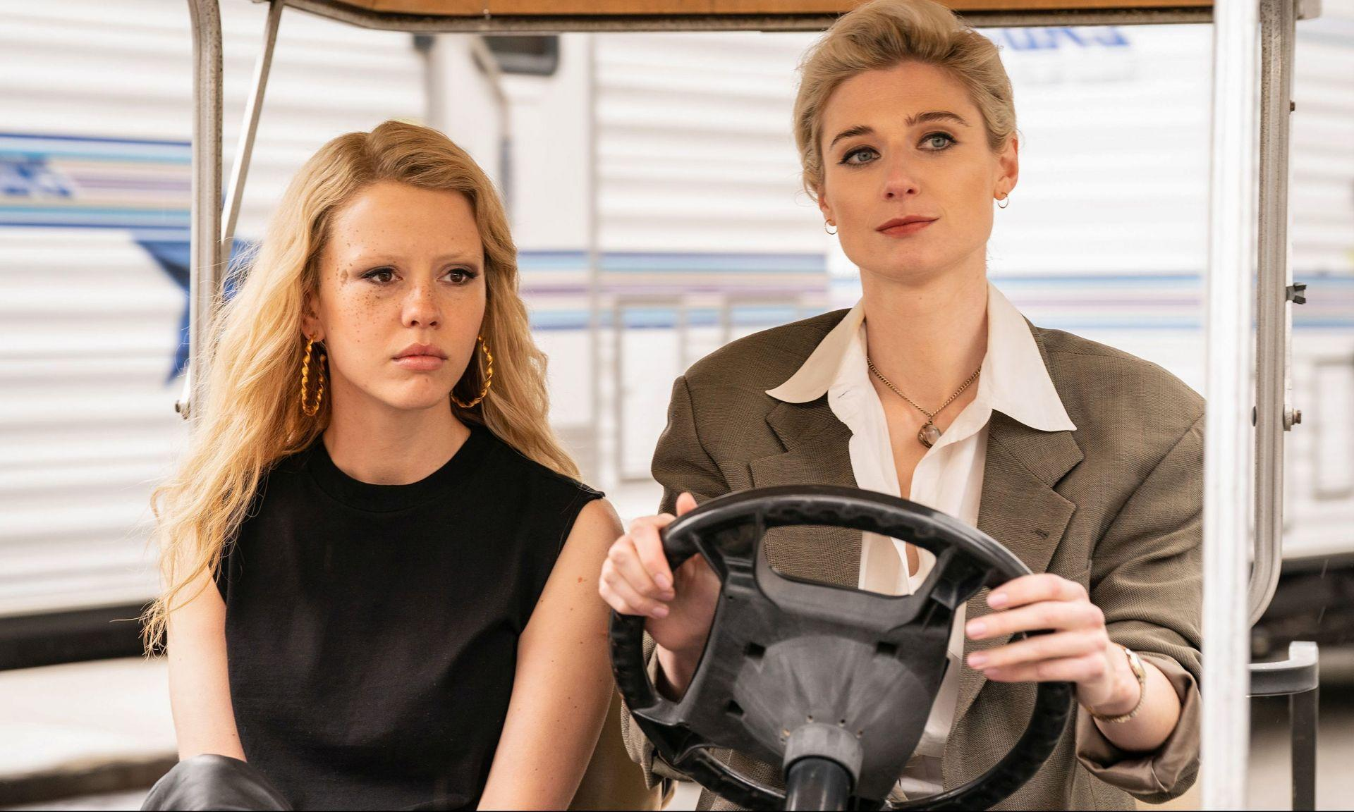 Zwei Frauen in einem Golfwagen. Eine fährt, während die andere zusieht. Beide haben blondes Haar und wirken ernst.