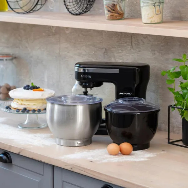 Scena kuchenna z czarnym mikserem, miskami, jajkami i ciastem. Półki z słoikami i drucianym koszem. Roślina po prawej.