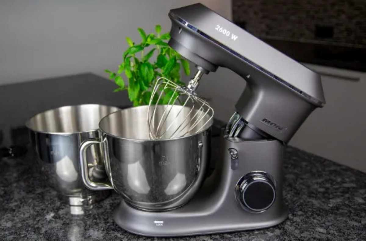 Szary mikser kuchenny z trzepaczką i dwiema miskami ze stali nierdzewnej stoi na granitowym blacie. Zielona roślina w tle.