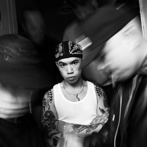 Mann in weißem Tanktop und Bandana mit Tätowierungen. Andere Personen sind im Hintergrund verschwommen.