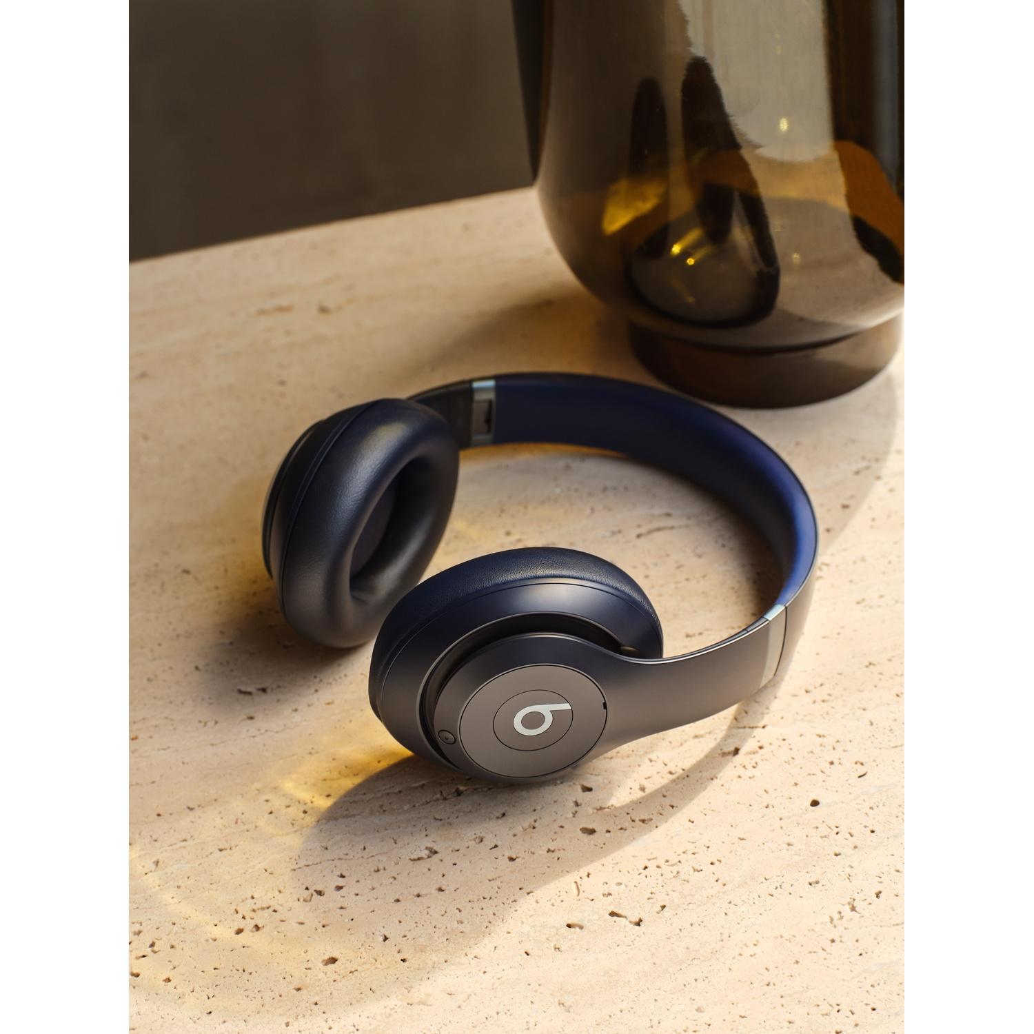 Cuffie Beats blu navy su una superficie beige strutturata, con un vaso di vetro scuro sullo sfondo. Le cuffie sono il punto focale.