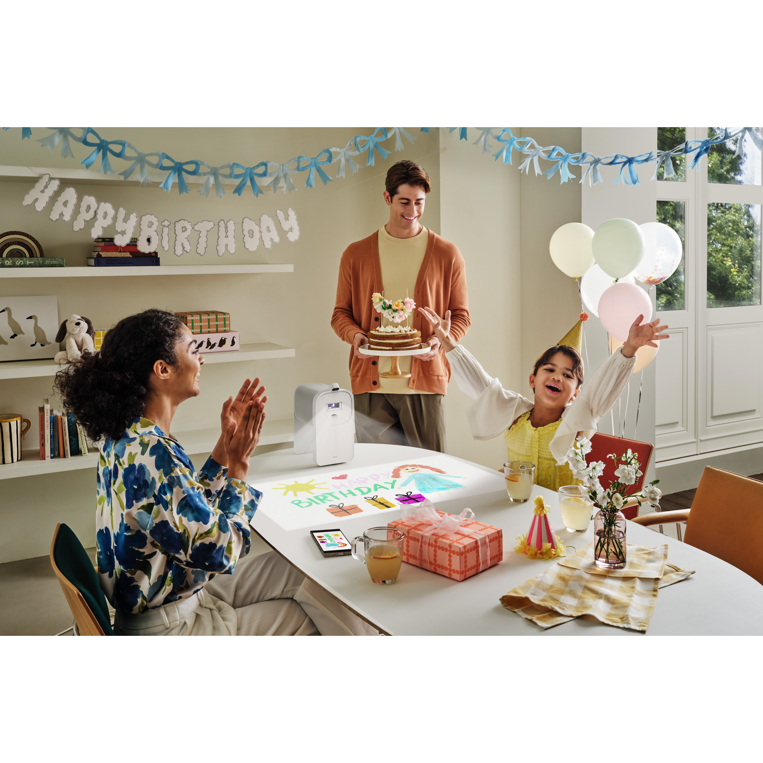 Une fête d'anniversaire avec un gâteau, des ballons et des gens heureux. Une femme applaudit, une fille fête et un homme tient le gâteau.