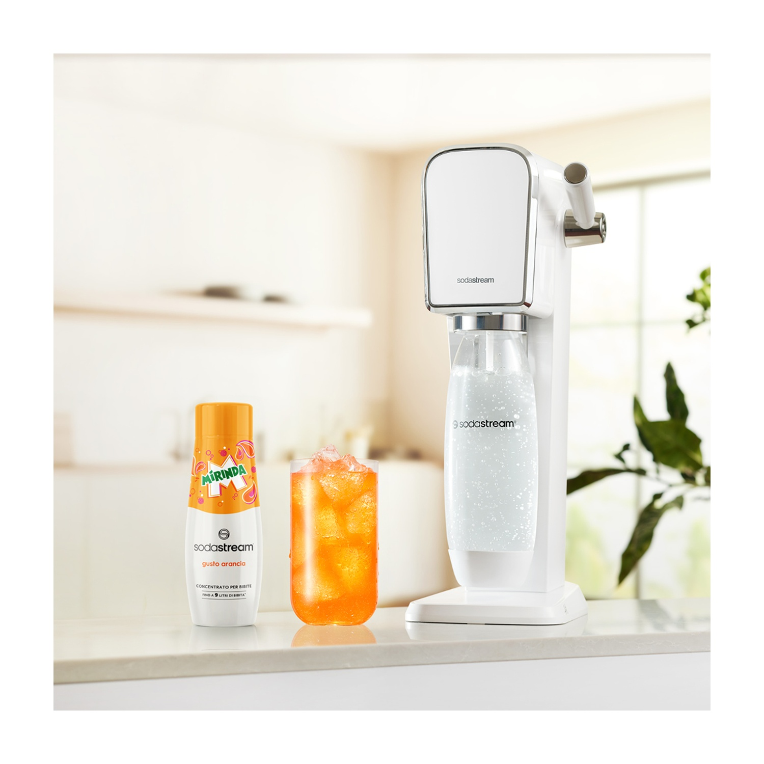 Una macchina Sodastream bianca, una bottiglia di concentrato di arancia Mirinda, un bicchiere di bevanda all'arancia con ghiaccio, il tutto su un bancone.