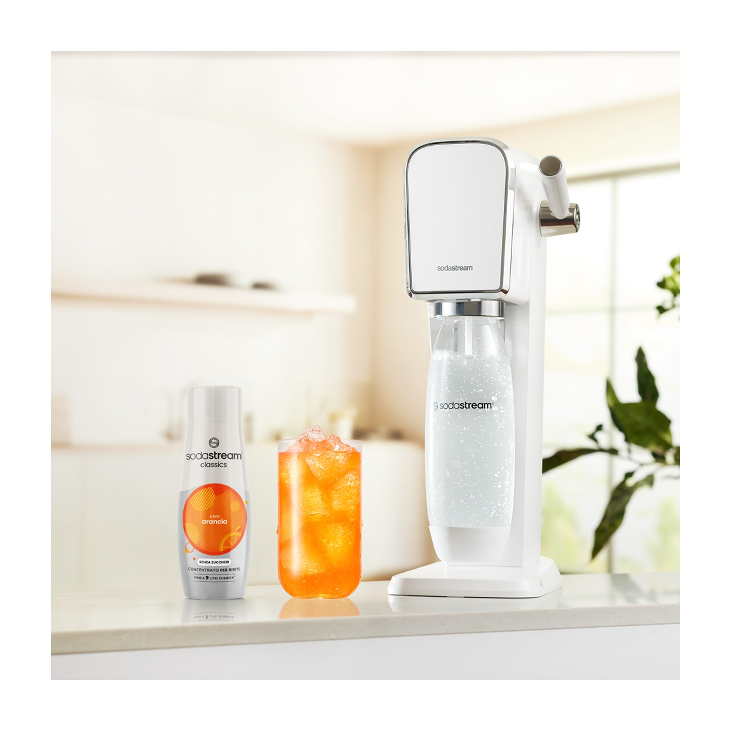 Una macchina SodaStream bianca, una bottiglia di sciroppo all'arancia e un bicchiere di bevanda all'arancia con cubetti di ghiaccio sono su un bancone.