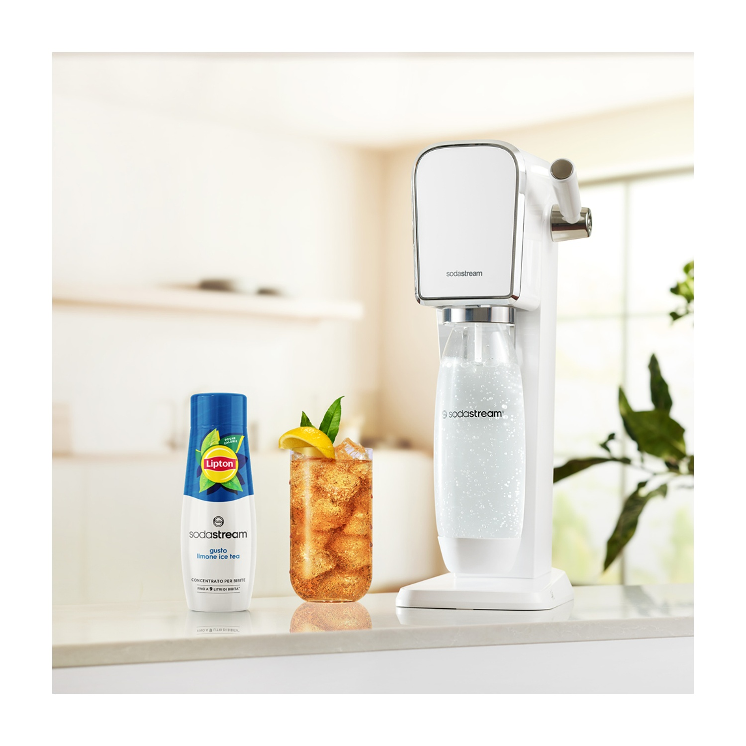 Una macchina SodaStream bianca, concentrato di tè freddo Lipton e un bicchiere di tè freddo al limone su una superficie bianca.