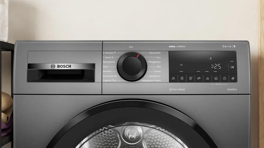 Un lave-linge Bosch gris avec un panneau de contrôle, un cadran et un affichage numérique indiquant 1:25. Fond mur beige.