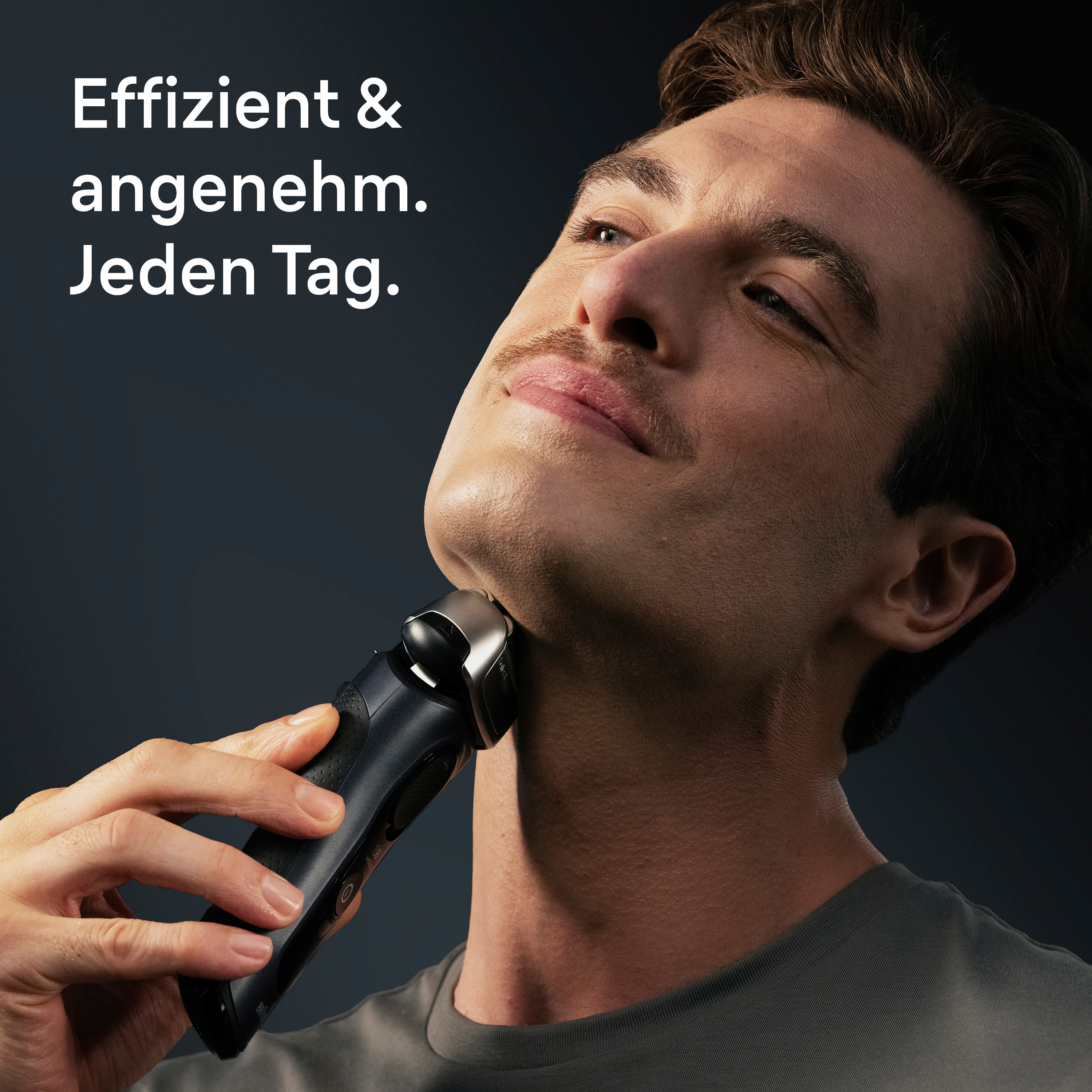 Ein Mann rasiert sich mit einem Elektrorasierer den Hals. Er hat einen Schnurrbart und trägt ein graues Hemd.