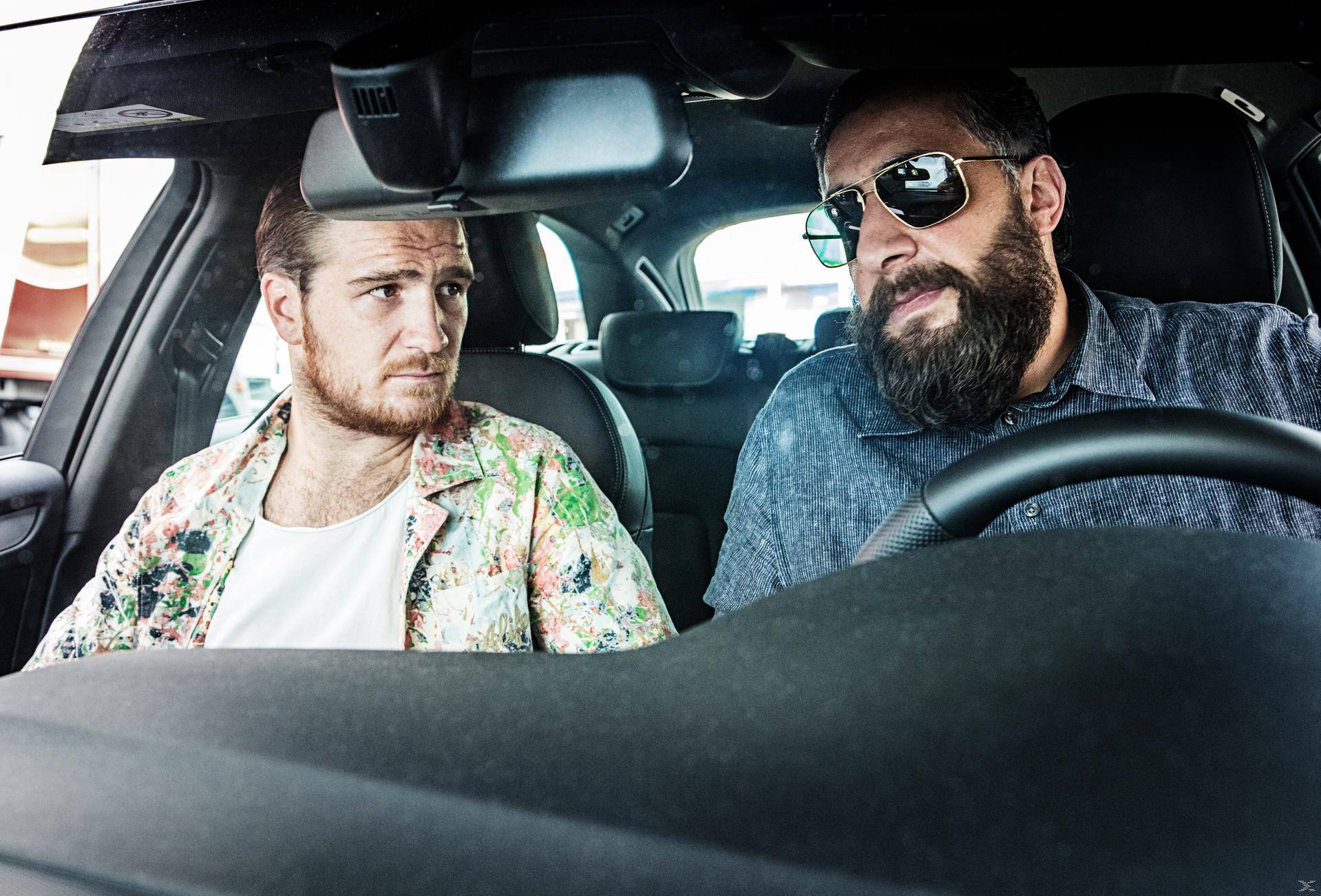 Zwei Männer in einem Auto. Einer fährt mit Sonnenbrille, der andere schaut ihn an. Beide haben Bärte.